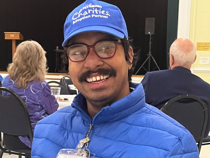 Smiling man with a mustache wearing a blue hat, blue jacket, name tag and glasses.