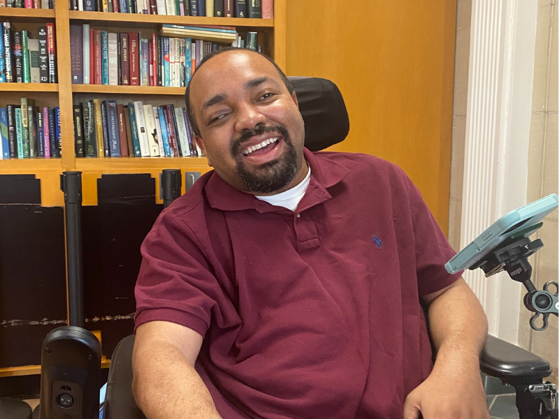 Smiling man seated in wheelchair in front of a bookcase.
