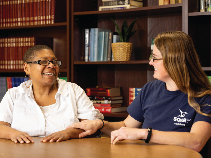 A program participant and a case manager sit and discuss wellness ideas at a table in front of a bookcase.