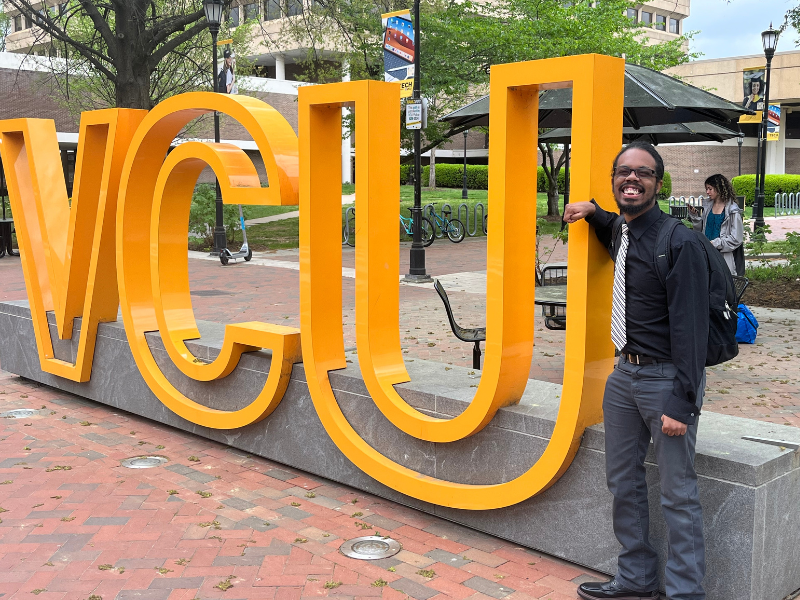 Troy Carter wears jeans, a button down shirt and a tie, posing next to a large VCU sign.