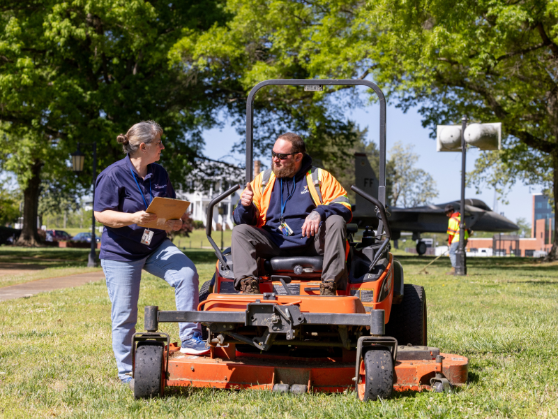 Woman holding a clipboard talkes to a man wearing a high visibility vest on a riding mower.