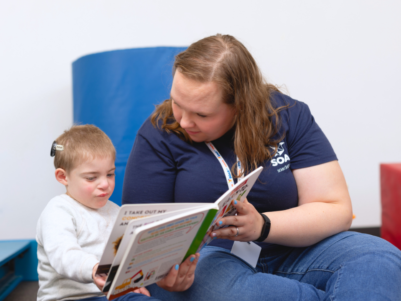 Woman wearing SOAR365 t-shirt is reading a book with a little boy who has cochlear implants.