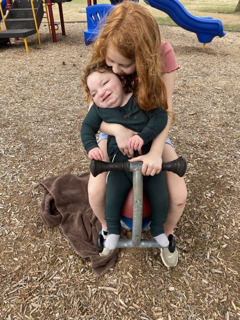 Anna is smiling as she sits on a piece of playground equipment, held by her sister.