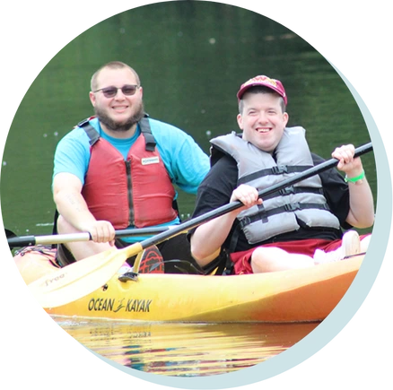 Two men kayaking on a calm lake, both smiling and wearing life vests during Summer Camp