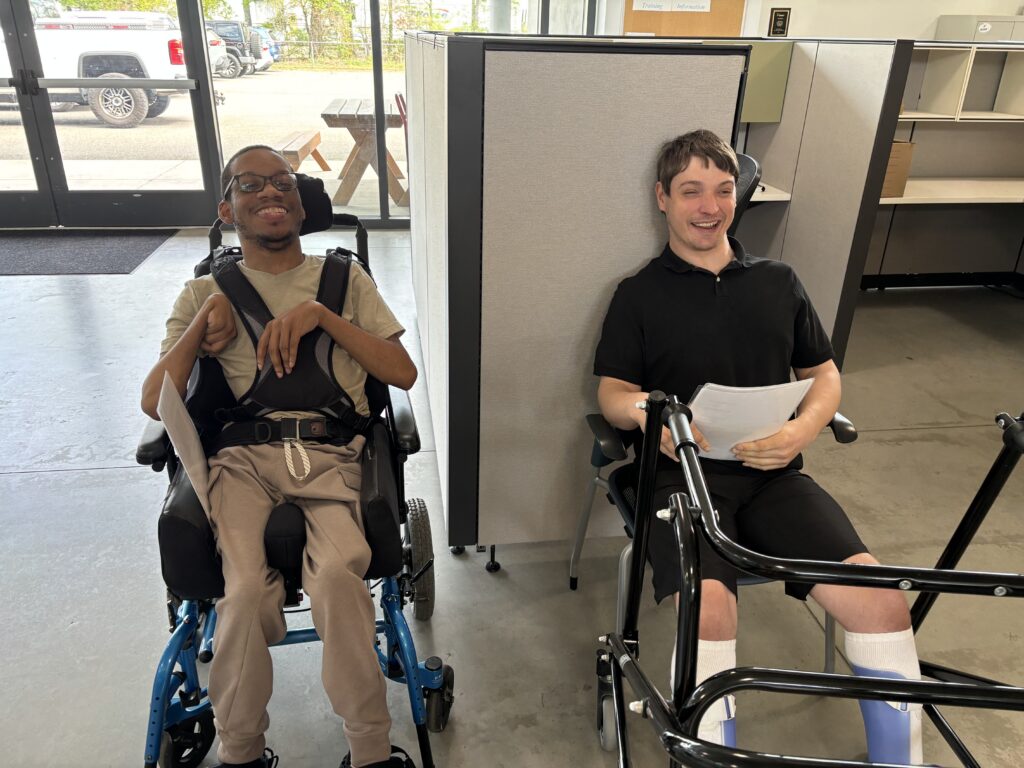 Rashad smiles as he sits in a wheelchair on the left, while Drew smiles and sits on a chair next to him with his walker nearby. Both are holding menus.