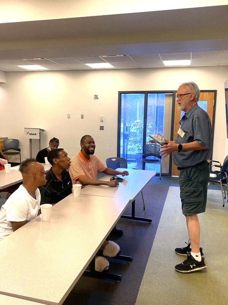 Tom stands before a seated group, discussing a book in his hand.
