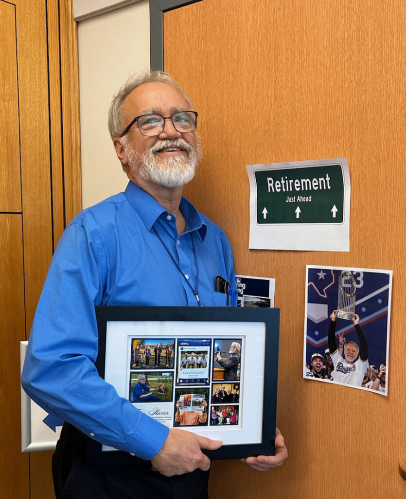 Tom holds a framed photo collage and stands smiling in front of a door with a sign that says "Retirement just ahead."